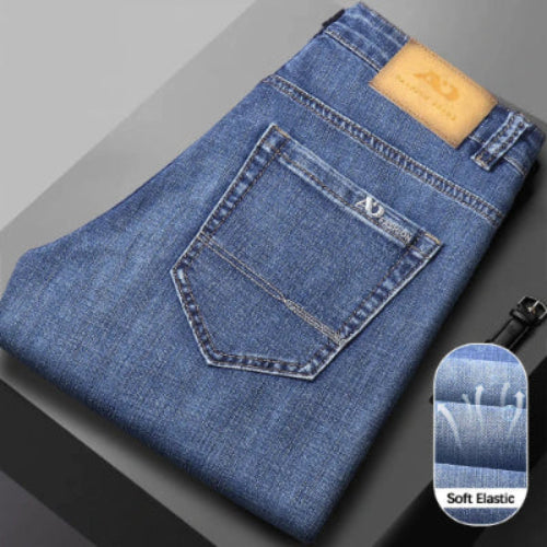 A folded pair of blue denim jeans shown on a dark gray surface, with the back pocket and a tan leather brand patch 