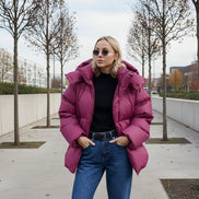 A woman wearing an oversized magenta puffer jacket, blue jeans, and sunglasses, standing against a beige stone wall.