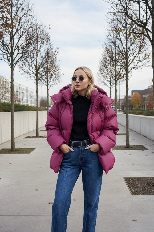 A woman wearing an oversized magenta puffer jacket, blue jeans, and sunglasses, standing against a beige stone wall.