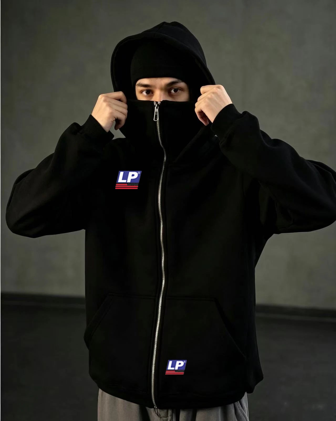 A person wearing a black zip-up hoodie with a silver zipper, a black balaclava, and a black hood, holding the hood up around their face. The hoodie features a white 'LP' logo patch on the left chest and a matching patch near the lower front pocket.