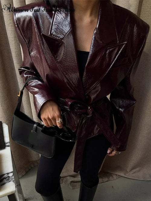 Close-up of a woman wearing a glossy dark brown jacket with wide lapels and a tied waist belt, holding a black handbag