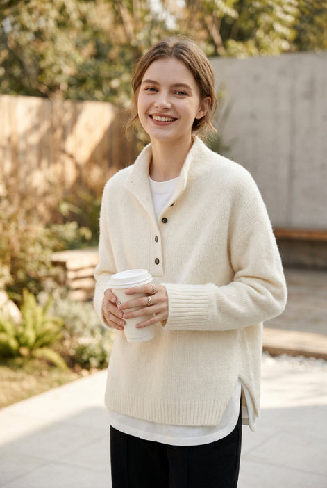 A woman wearing a cream-colored knit cardigan sweater with a high collar and button details, holding a white coffee cup in a bright indoor setting.