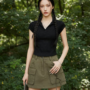 A model wearing a black short-sleeve knit polo shirt tucked into olive green cargo pants, accessorized with a beige cap, layered gold necklaces, and double braids.