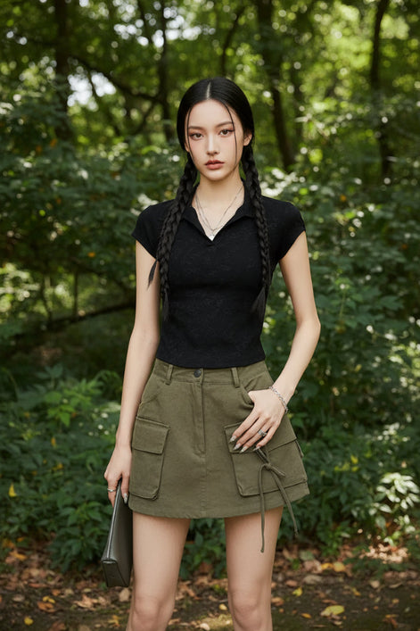 A model wearing a black short-sleeve knit polo shirt tucked into olive green cargo pants, accessorized with a beige cap, layered gold necklaces, and double braids.