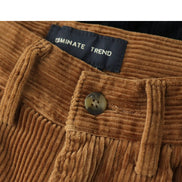 Close-up of brown corduroy trousers showing the waistband, belt loops, button, and a black label that reads OMINATE TREND.