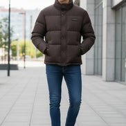 A dark brown puffer jacket with a high collar and snap-button front, shown on a hanger with a hand holding the lower right side. Three feature callout graphics on the left mention 85% down, thick warm lining, and fluffy soft texture.