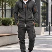 A man wearing a dark gray zip-up hoodie and matching cargo sweatpants with orange zipper accents, 