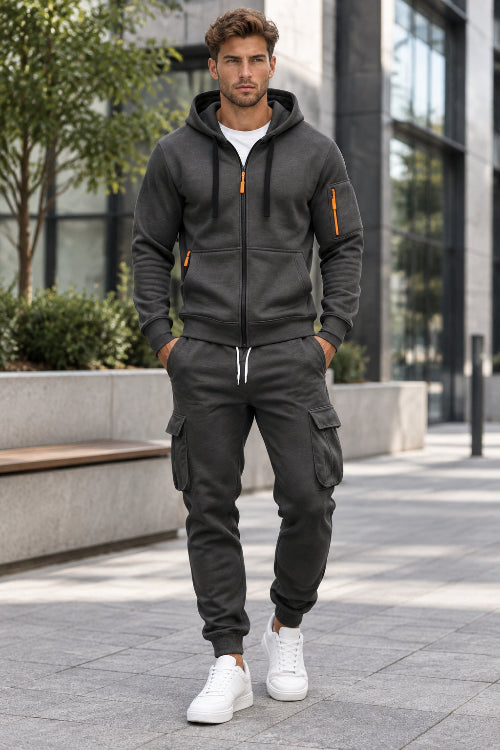 A man wearing a dark gray zip-up hoodie and matching cargo sweatpants with orange zipper accents, 
