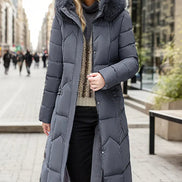A woman wearing a long gray puffer coat with a large faux-fur trimmed hood and quilted chevron pattern, standing on a city street. The brand name VIGOJANY 