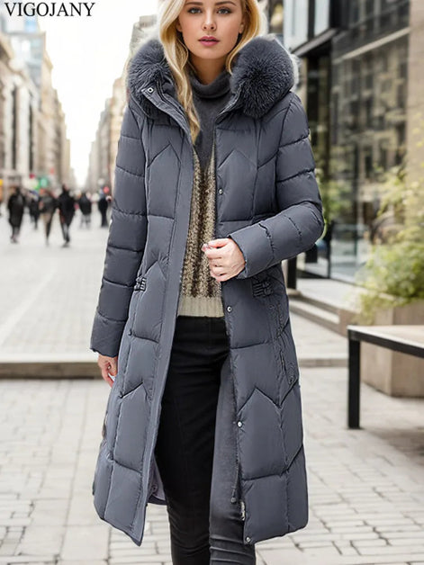 A woman wearing a long gray puffer coat with a large faux-fur trimmed hood and quilted chevron pattern, standing on a city street. The brand name VIGOJANY 