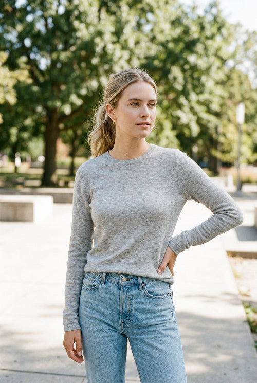 A woman wearing a fitted light gray long-sleeve turtleneck sweater and light blue jeans, shown in a mirror selfie against a beige tiled wall.