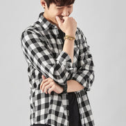 A man wearing a black and white checkered long-sleeve button-up shirt over a black undershirt and blue jeans, standing against a light gray background. 