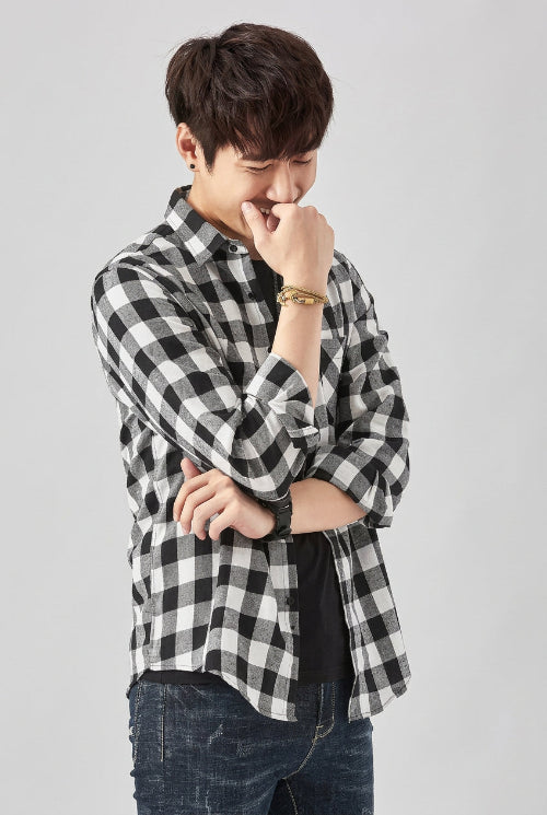 A man wearing a black and white checkered long-sleeve button-up shirt over a black undershirt and blue jeans, standing against a light gray background. 