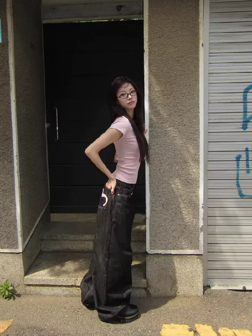 Side view of a woman wearing dark-wash loose-fit jeans with a pink heart outline on a back pocket, standing outside by steps.