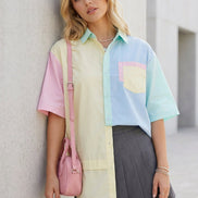 A woman wearing a pastel multicolor short-sleeve shirt with pink, light blue, and cream panels, a dark gray pleated skirt, and a white face mask, standing in a doorway.