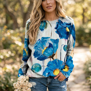A woman wearing a long-sleeve crewneck sweatshirt with a vibrant blue and yellow abstract floral print on a white background, paired with distressed blue jeans.