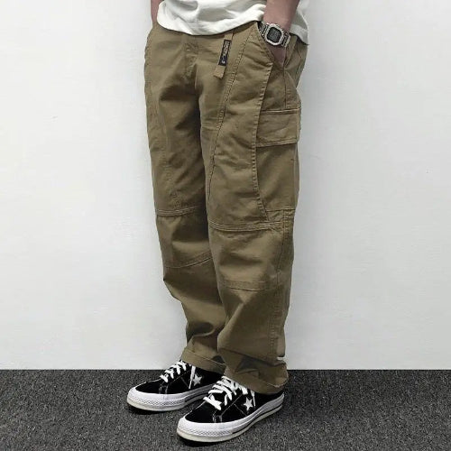Model wearing khaki loose-fit cargo pants with large flap thigh pockets and stitched knee panels, paired with black sneakers.