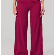 A model is shown from the waist down wearing high-waisted, wide-leg athletic pants in a deep magenta color, paired with white sneakers, against a light gray studio background.