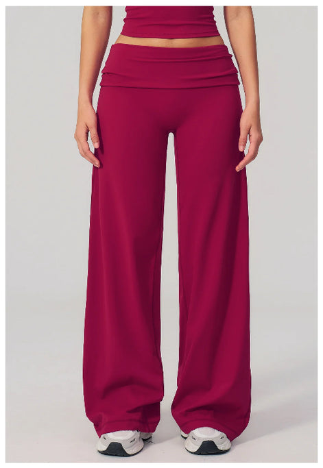 A model is shown from the waist down wearing high-waisted, wide-leg athletic pants in a deep magenta color, paired with white sneakers, against a light gray studio background.