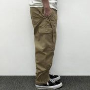 Side view of a model wearing khaki loose-fit cargo pants with a large flap thigh pocket, back pocket, and stitched knee panels, paired with black sneakers.