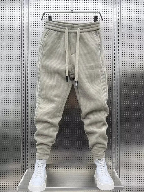 Beige baggy sweatpants hanging on a hanger against a perforated metal wall, with white sneakers below.