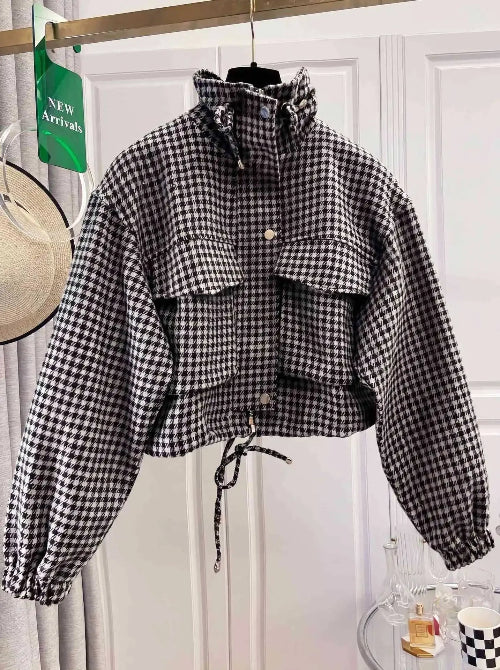 Black and white houndstooth utility jacket on a hanger, showing a raised drawcord collar, chest flap pockets, front snap buttons, elasticated cuffs and a tied drawcord hem