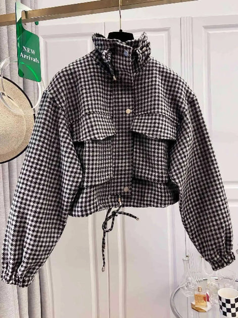 Black and white houndstooth utility jacket on a hanger, showing a raised drawcord collar, chest flap pockets, front snap buttons, elasticated cuffs and a tied drawcord hem