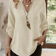Woman wearing a beige blouse with a necklace in a room with a window.