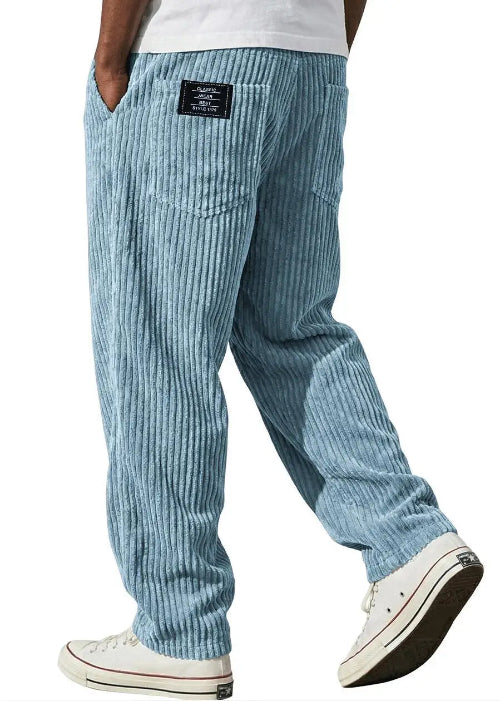 Light blue striped corduroy pants shown from the back on a model, featuring a black brand patch on the back pocket and white low-top sneakers.