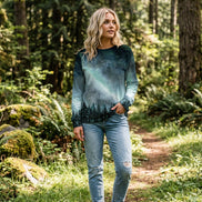 A woman wearing a long-sleeve crewneck sweatshirt with a galaxy night sky print, featuring a green aurora borealis glow and a dark pine forest silhouette at the bottom, paired with blue jeans.