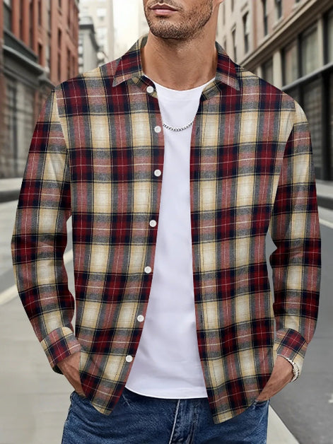 A man wearing a long-sleeve button-up plaid flannel shirt in red, beige, and black over a white t-shirt and blue jeans, standing on a city street.