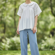 A white short-sleeve blouse with a Peter Pan collar, gathered elastic waist, and pleated details, hanging on a clear hanger against a textured gray wall.