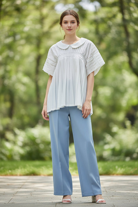 A white short-sleeve blouse with a Peter Pan collar, gathered elastic waist, and pleated details, hanging on a clear hanger against a textured gray wall.