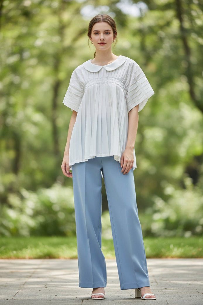 A white short-sleeve blouse with a Peter Pan collar, gathered elastic waist, and pleated details, hanging on a clear hanger against a textured gray wall.