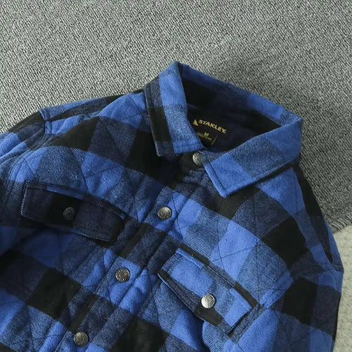 A close-up of a blue and black plaid flannel shirt by Stanley, featuring a quilted texture, button-down front, and chest pockets, laid flat on a gray textured surface.