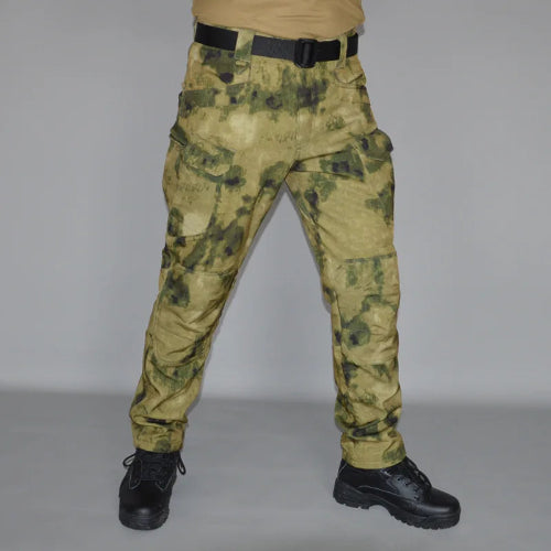 Front view of a model wearing tan and olive green camouflage cargo pants with a black belt and black lace-up boots against a gray background.