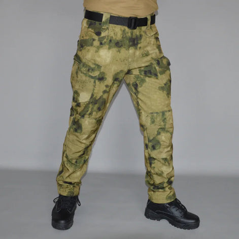 Front view of a model wearing tan and olive green camouflage cargo pants with a black belt and black lace-up boots against a gray background.