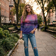 A woman wearing a long-sleeve crewneck sweatshirt with a purple and pink watercolor sunset print, paired with blue jeans and holding a small bouquet of white roses.