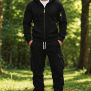 A man wearing a black zip-up hoodie and matching black jogger pants with cargo pockets, standing in a sunlit forest clearing.