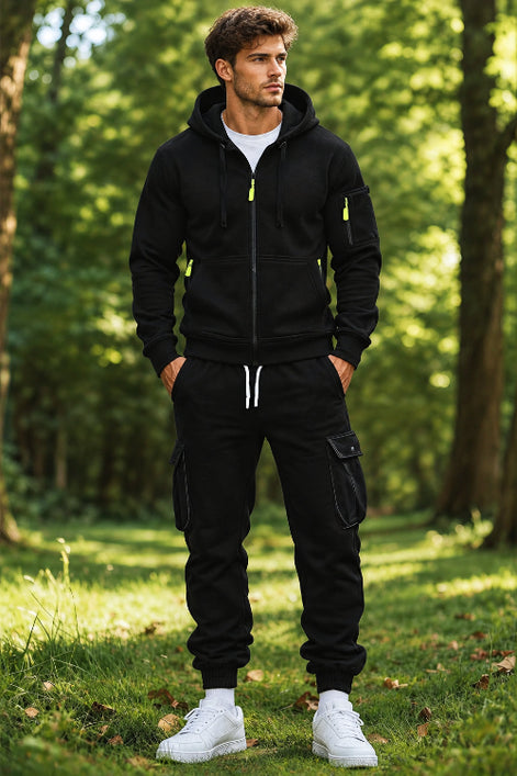 A man wearing a black zip-up hoodie and matching black jogger pants with cargo pockets, standing in a sunlit forest clearing.