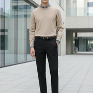 A person wearing slim black trousers with a gray knit sweater and black loafers, standing indoors in front of a modern glass-paneled wall.