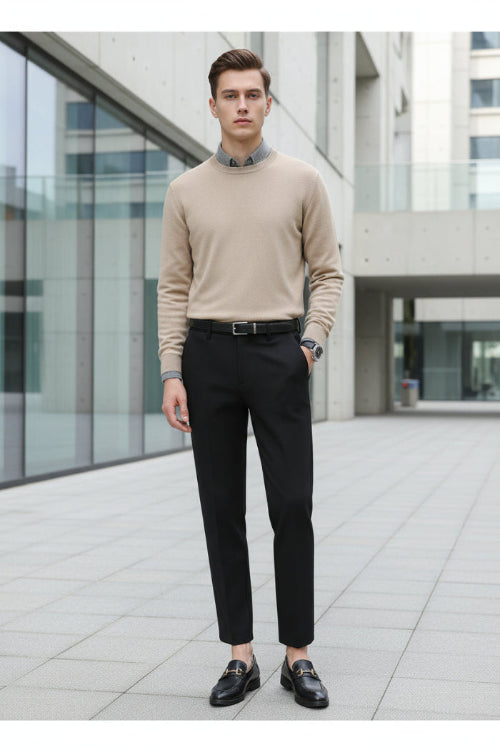 A person wearing slim black trousers with a gray knit sweater and black loafers, standing indoors in front of a modern glass-paneled wall.