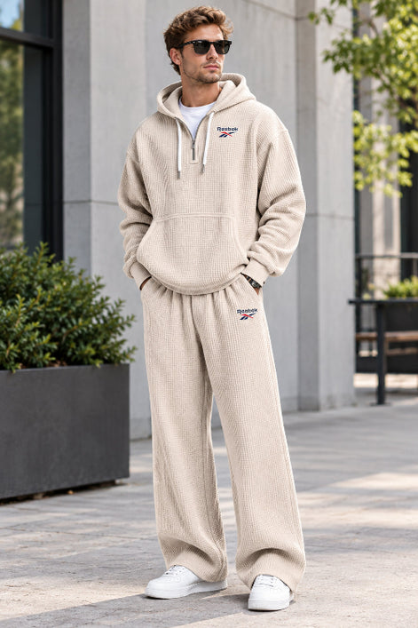 A man wearing a beige waffle-knit Reebok tracksuit, featuring a pullover hoodie with a drawstring hood and matching sweatpants, 