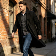 A man walking down a sunlit city street wearing a long black overcoat over a dark button-up shirt and blue jeans.