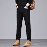 A person wearing slim black pants, a black long-sleeve top, tan lace-up boots, and carrying a brown shoulder bag, photographed against a plain light gray studio background.