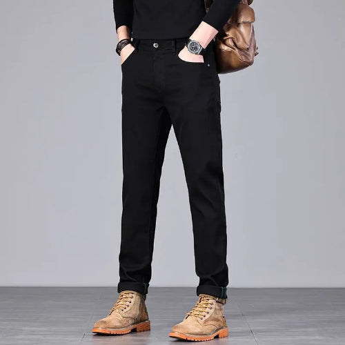 A person wearing slim black pants, a black long-sleeve top, tan lace-up boots, and carrying a brown shoulder bag, photographed against a plain light gray studio background.