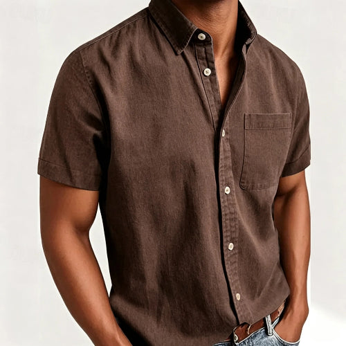 A man wearing a brown short-sleeve button-up shirt with a chest pocket, paired with blue jeans and a brown leather belt, shown against a plain light background.