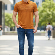 A close-up of a men's short-sleeve polo shirt in a warm orange-brown color, held by a hand to show its texture. The shirt features a subtle landscape pattern on the front and a small embroidered logo on the chest.