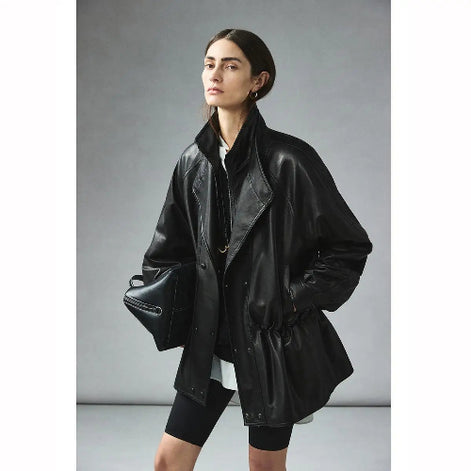 Woman wearing a black faux leather jacket with layered collars and a gathered waist, holding a black handbag against a gray background⠋