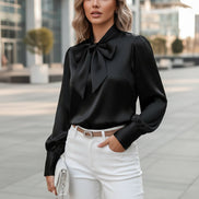 Black satin blouse with a large bow tie worn by a person, sitting on a white chair.